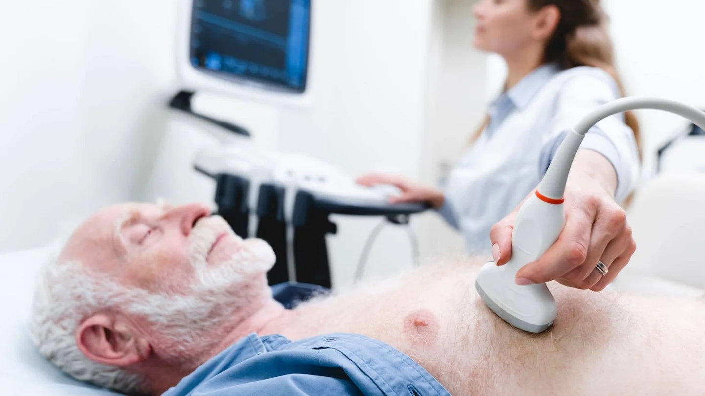 Ein &auml;lterer Mann liegt mit geschlossenen Augen auf einer Liege. Im Hintergrund ist eine &Auml;rztin zu sehen. Sie untersucht das Herz des Mannes per Ultraschall.