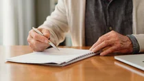 Eine &auml;ltere Person sitzt am Tisch und unterschreibt ein Dokument.
