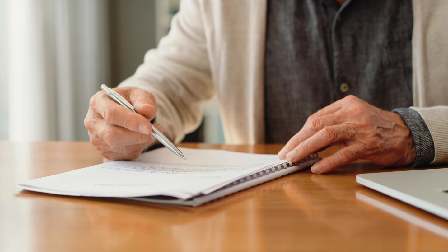 Eine &auml;ltere Person sitzt am Tisch und unterschreibt ein Dokument.