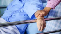 Eine Person liegt in einem Bett mit einem Bettgitter aus Metall. Sie hat den Arm auf das Bettgitter gelegt. Eine weitere Person ber&uuml;hrt diesen Arm.