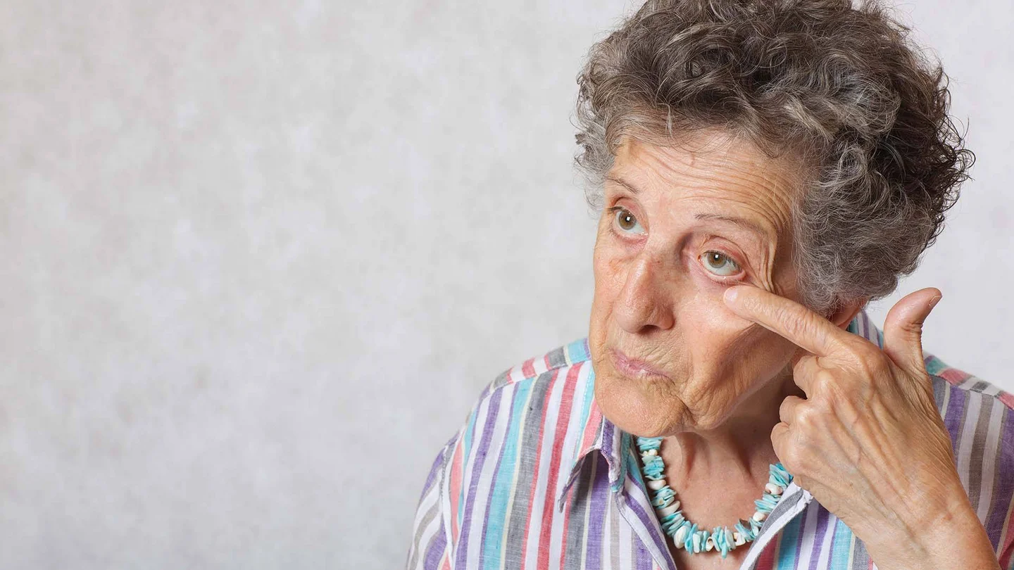 Eine &auml;ltere Dame fasst sich pr&uuml;fend ans Auge, als w&uuml;rde sie sich im Spiegel betrachten.
