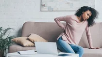 Eine Frau sitzt auf einer Couch. Mit einem Arm st&uuml;tzt sich die Frau ab, mit der anderen Hand fasst sie sich an den R&uuml;cken.