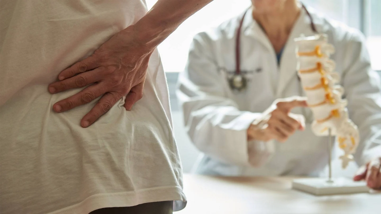 Frau greift sich an den unteren R&uuml;cken, ihr gegen&uuml;ber sitzt ein Arzt mit Wirbels&auml;ulenmodell.