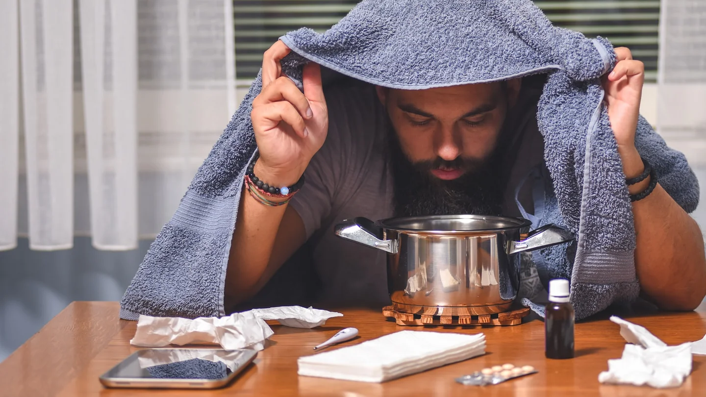 Ein Mann sitzt an einem Tisch, auf dem Taschent&uuml;cher, Fieberthermometer, Tabletten, Nasentropfen und ein Topf stehen. Der Mann h&auml;lt ein Handtuch &uuml;ber seinen Kopf und beugt sich &uuml;ber den Topf.