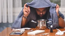 Ein Mann sitzt an einem Tisch, auf dem Taschent&uuml;cher, Fieberthermometer, Tabletten, Nasentropfen und ein Topf stehen. Der Mann h&auml;lt ein Handtuch &uuml;ber seinen Kopf und beugt sich &uuml;ber den Topf.