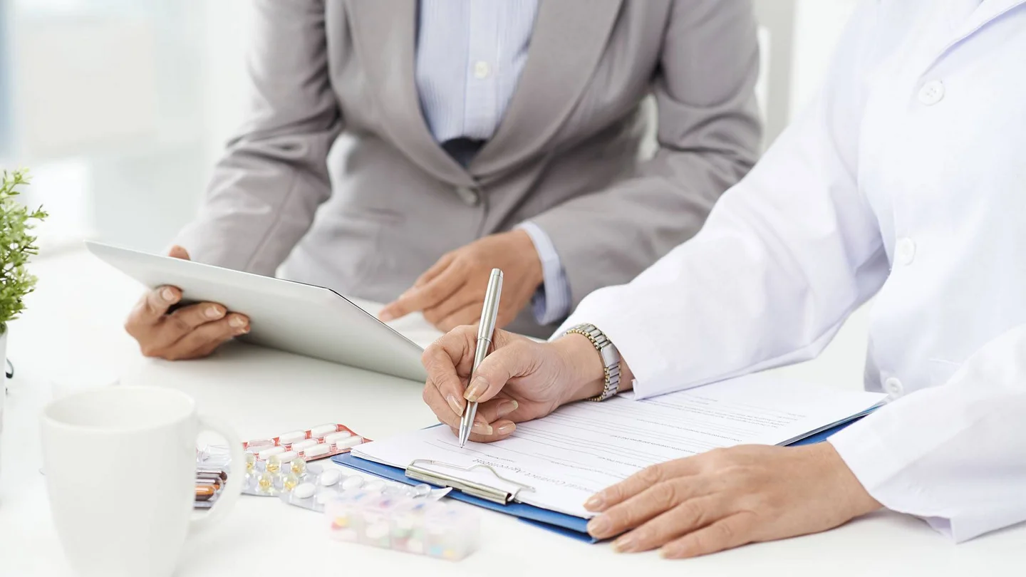 Eine Person im wei&szlig;en Kittel f&uuml;llt ein Dokument aus. Daneben sitzt eine Person, die auf ein Tablet zeigt.