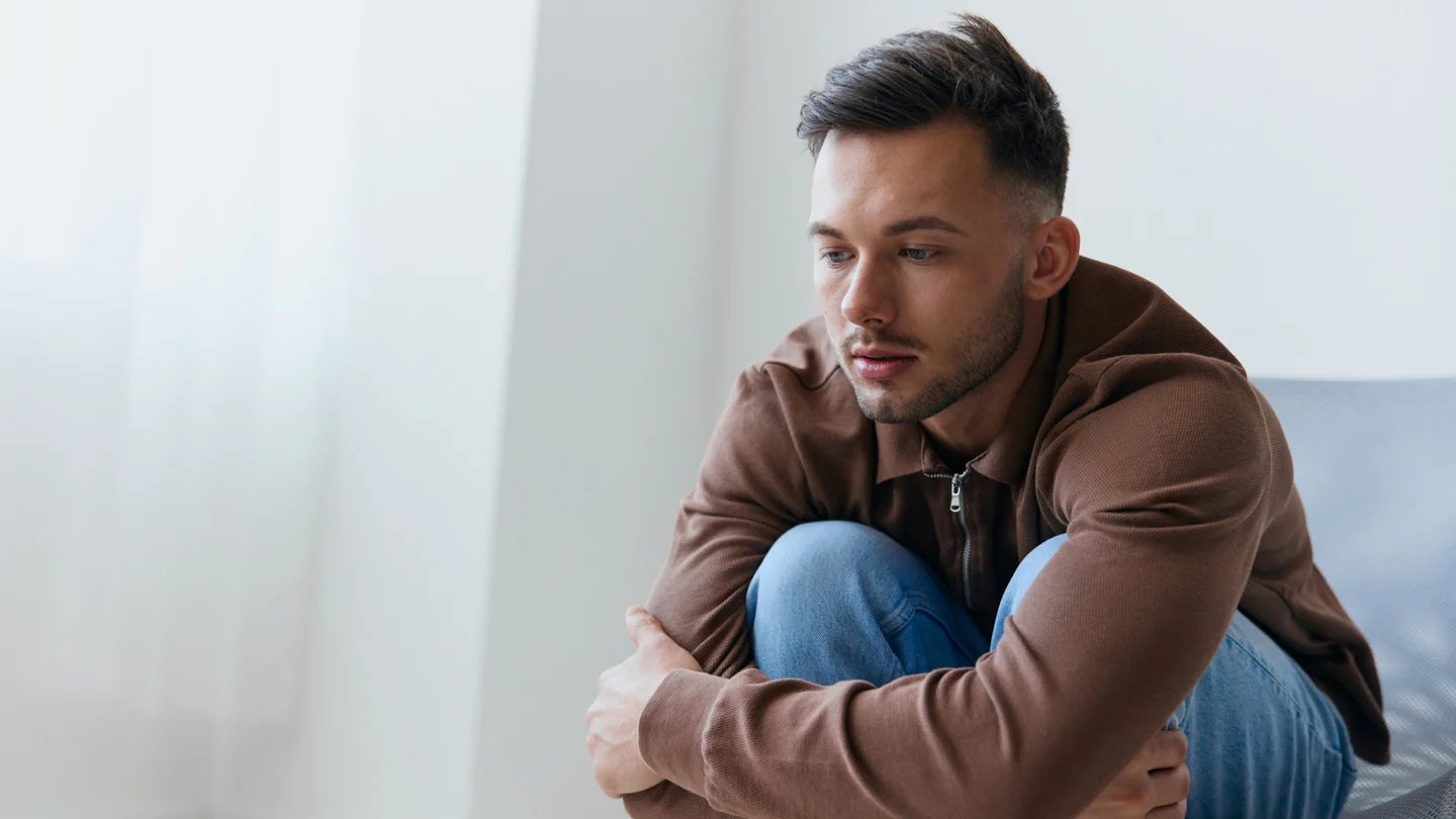 Eine junger Mann sitzt vorgebeugt auf einem Stuhl und hat die Arme um seine Knie geschlungen.