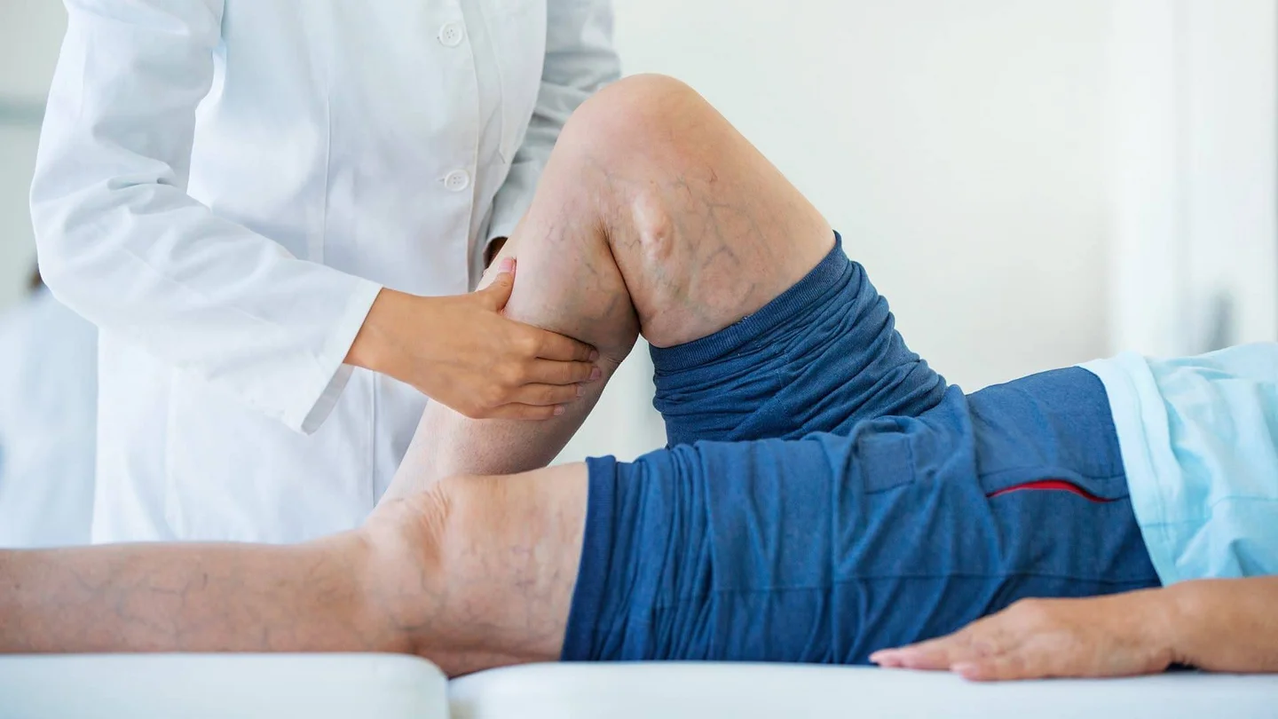 Eine Person im wei&szlig;en Kittel tastet die aufgestellte Wade eines Patienten ab, der auf einer Behandlungsliege liegt.