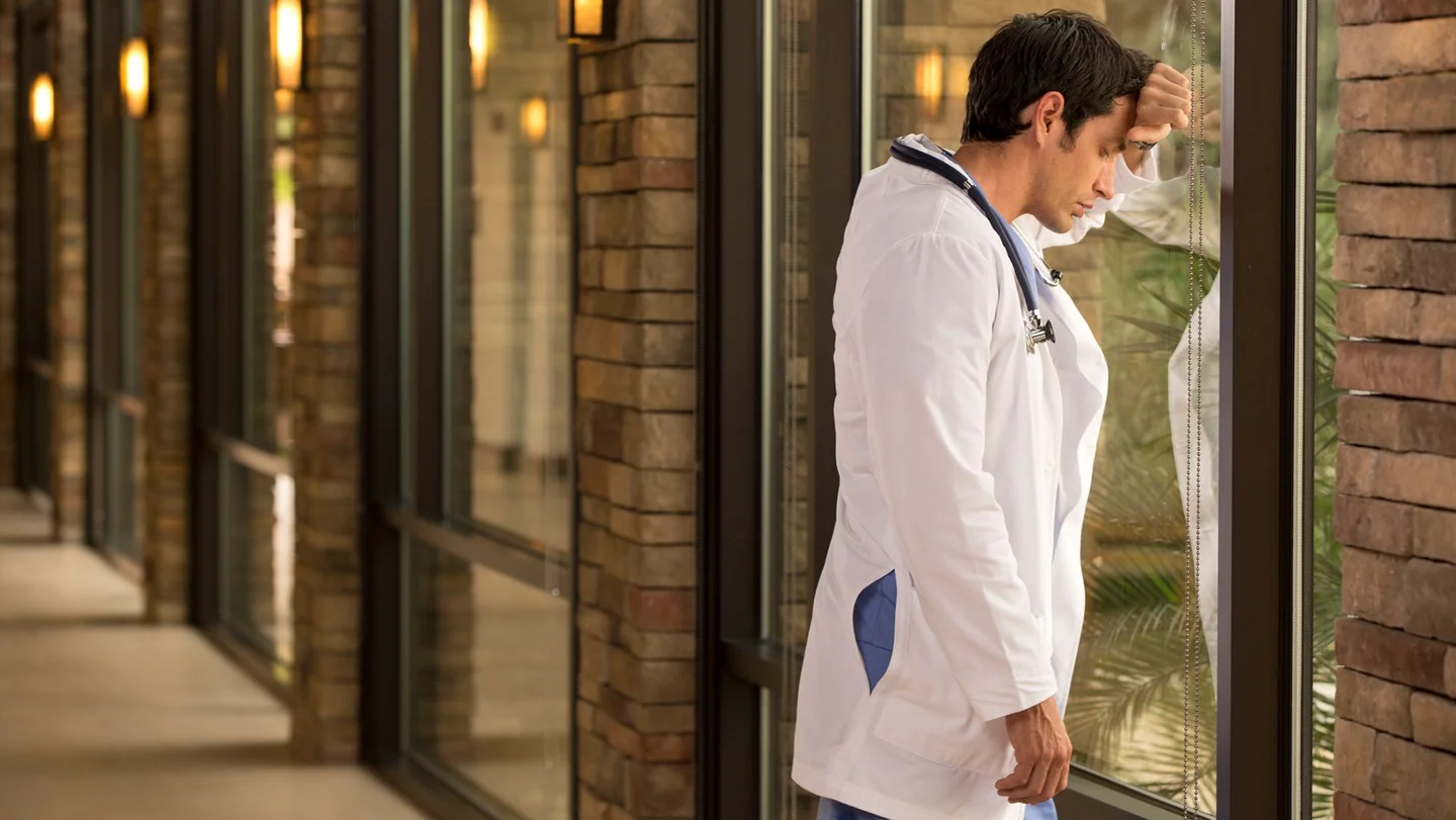 Ein junger Arzt steht mit gesenktem Kopf am Fenster. 