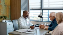 Chronische lymphatische Leukämie (CLL): Ein Arzt sitzt an seinem Schreibtisch und führt ein Gespräch mit einem älterern Mann und einer Frau.