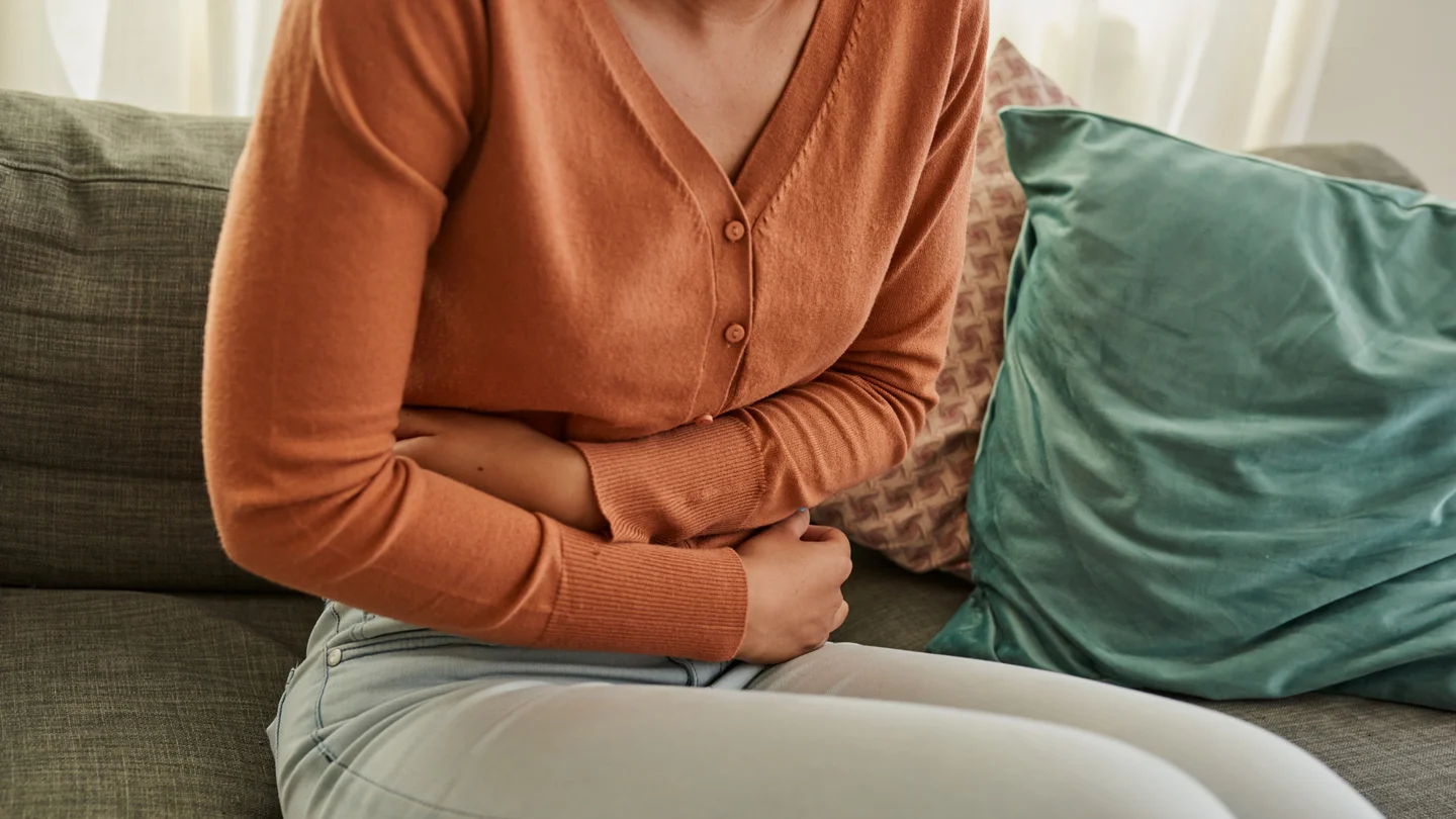 Hepatitis A: Eine Frau sitzt auf einem Sofa und hält die Arme über dem Bauch. Sie hat Bauchschmerzen.