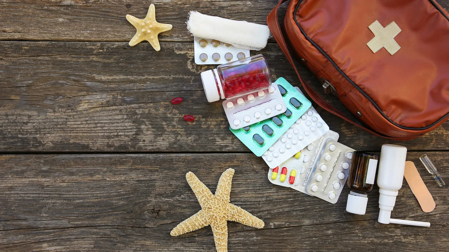 Auf verwitterten Holzdielen liegen neben zwei Seesternen Arzneimittel, Tablettenblister, und Verbandmaterial aus einer Reise-Medikamententasche.