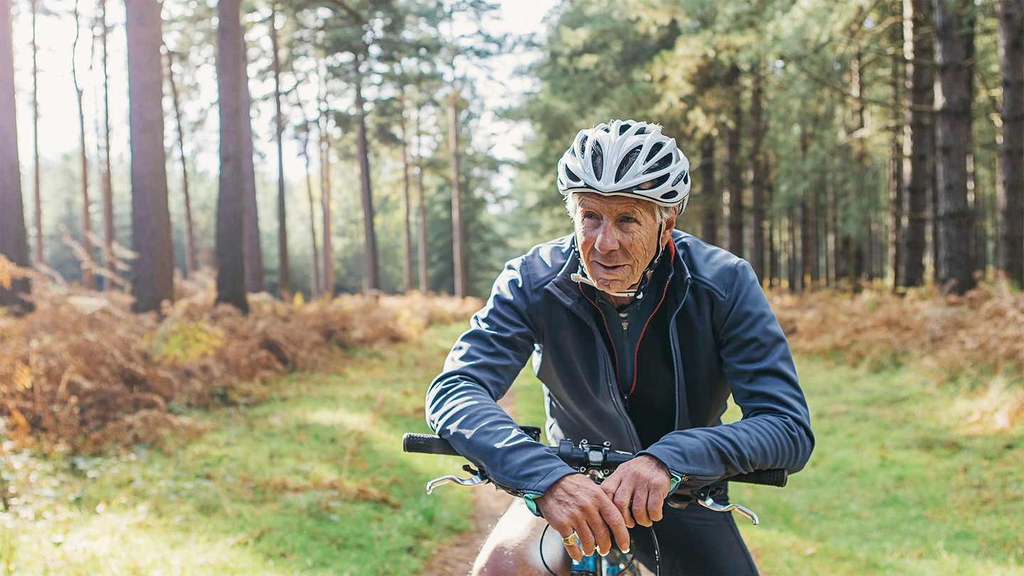 &Auml;lterer Mann in Fahrradkleidung im Wald st&uuml;tzt sich auf den Lenker seines Fahrrads.